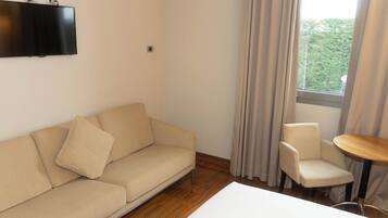 Ein Zimmer mit einem beigefarbenen Sofa, einem kleinen runden Tisch und einem an der Wand befestigten Fernseher.