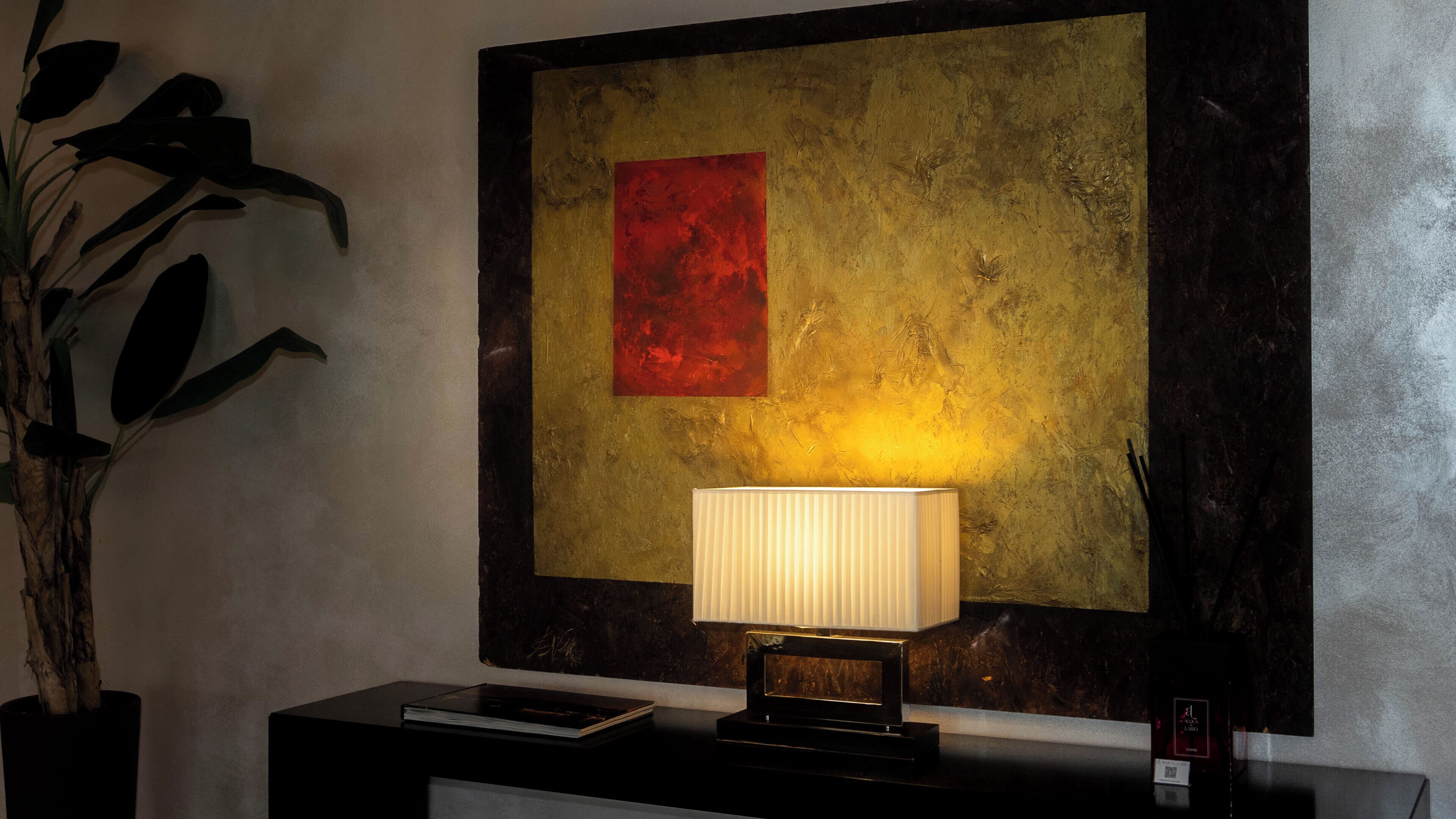 A room with a framed abstract painting, a standing lamp, a potted plant, and a book on a side table.