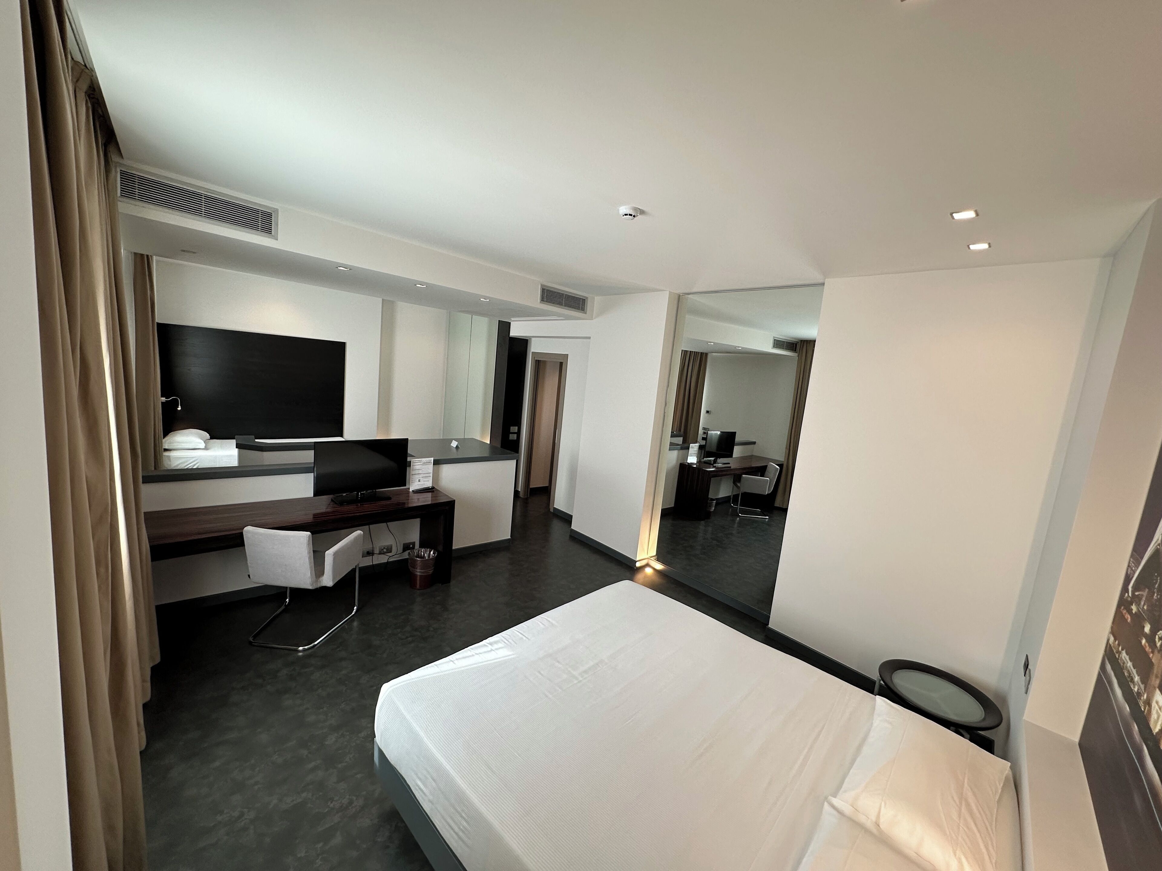 A modern hotel room with a large bed, a desk with a computer, a TV, and a mirror.