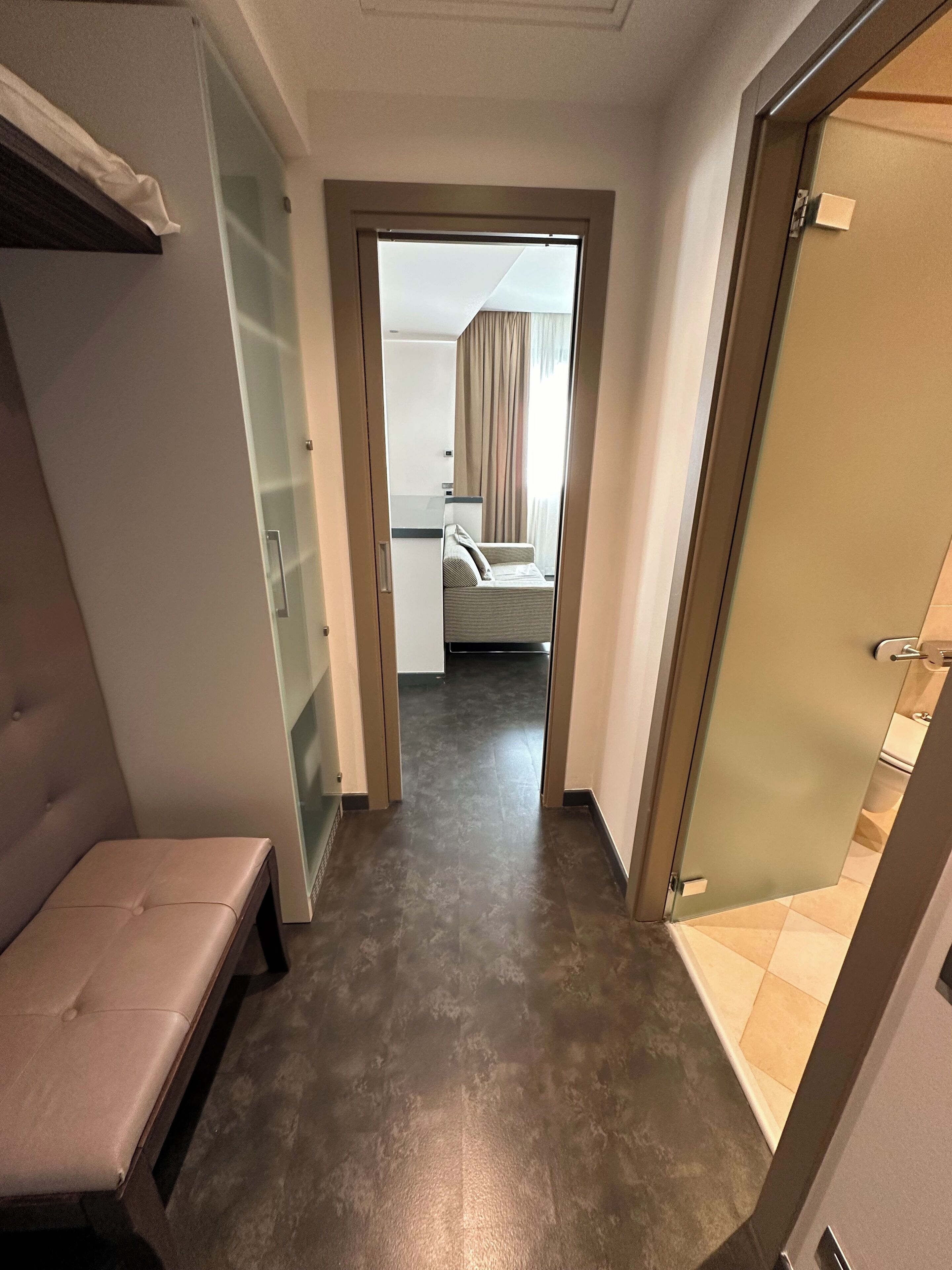 A hotel room with a bed, a bench, a glass door, and a view into another room.