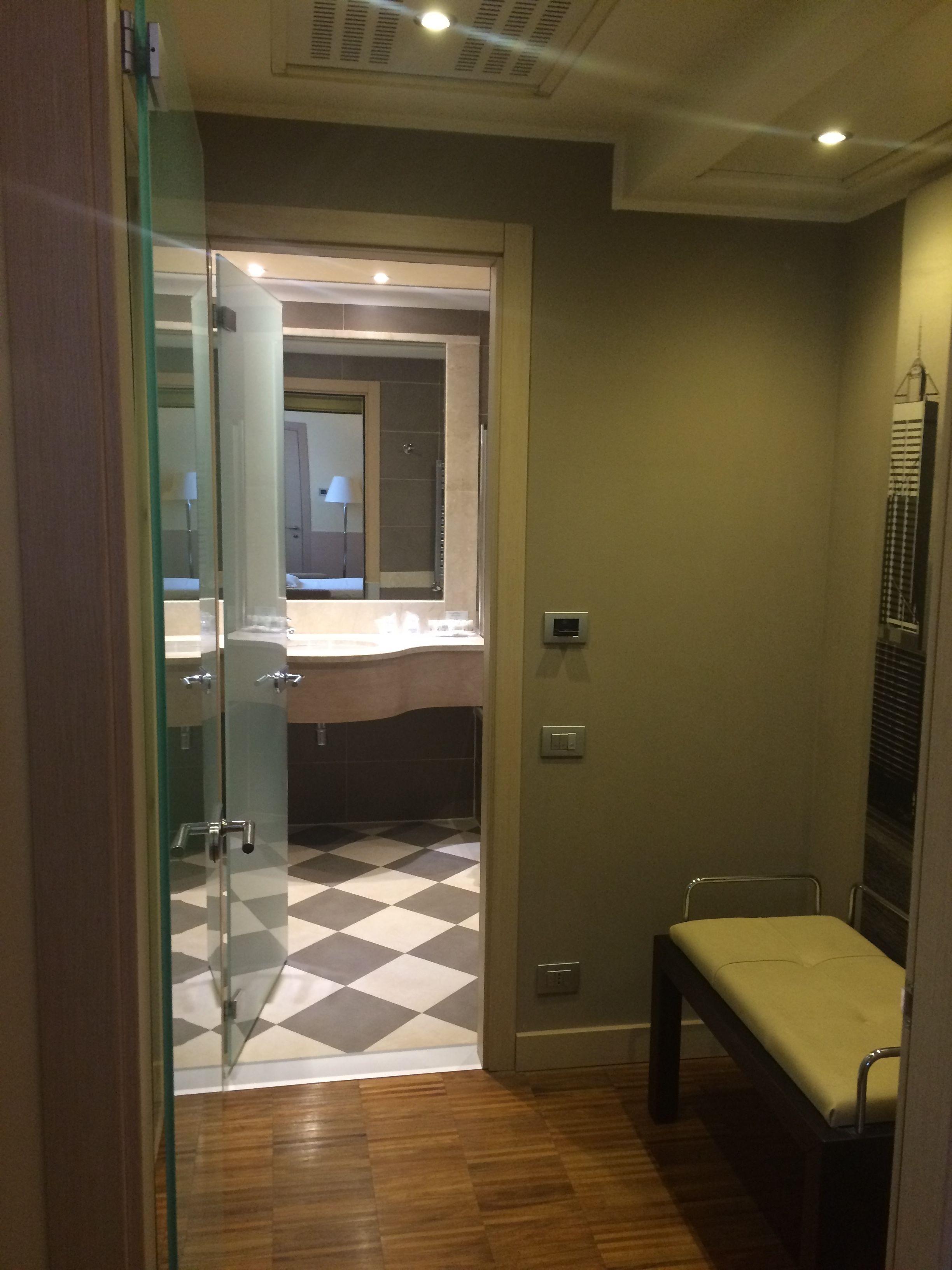 A hotel room with a checkered floor, a bed, a bench, a mirror, and a door leading to a bathroom.