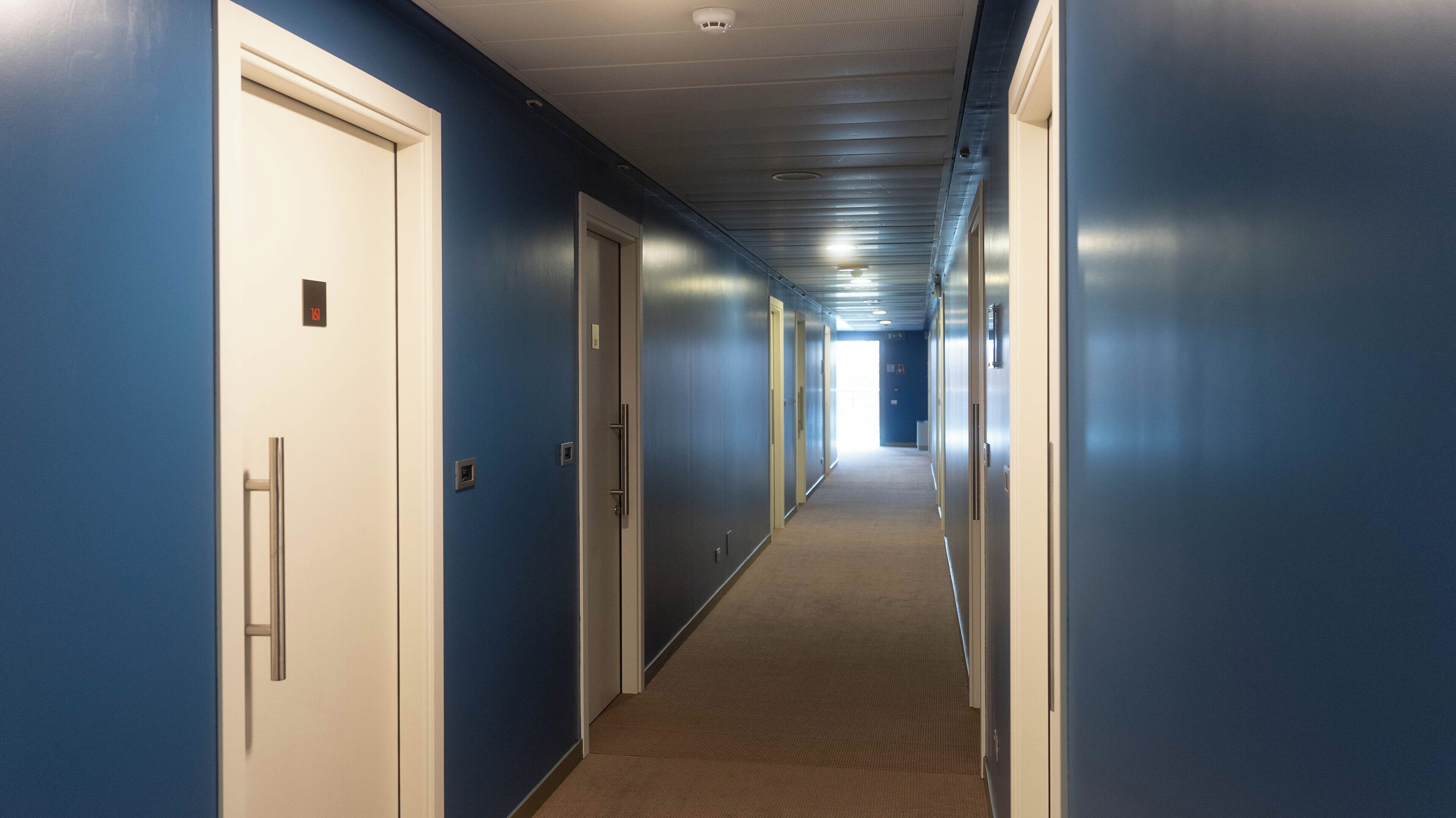 Un long couloir étroit aux murs bleus et aux portes blanches, qui mène à une pièce lumineuse au bout.