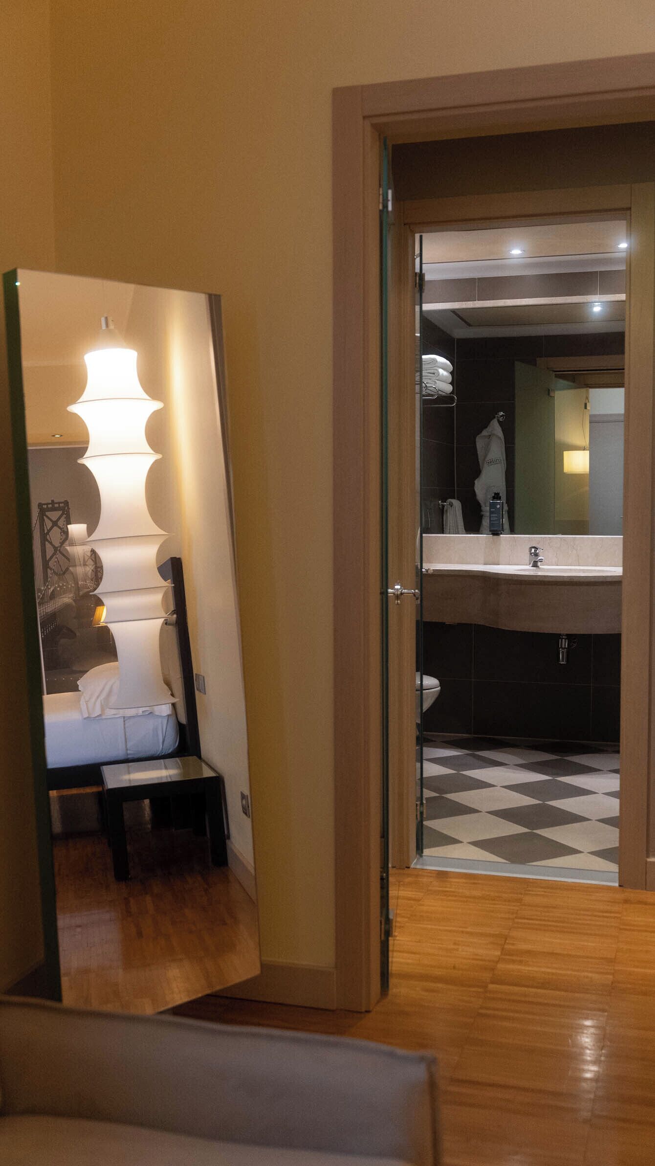 Habitación de hotel con suelo de madera, una cama con ropa de cama blanca, una mesita de noche, un espejo grande con una lámpara de diseño y un baño visible a través de una puerta abierta.