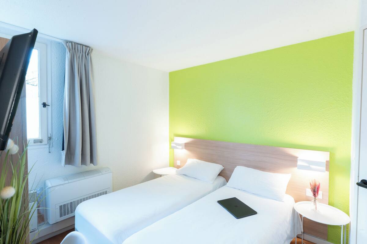 A hotel room with two beds, a window with curtains, an air conditioner, a bedside table with a lamp, and a green accent wall.