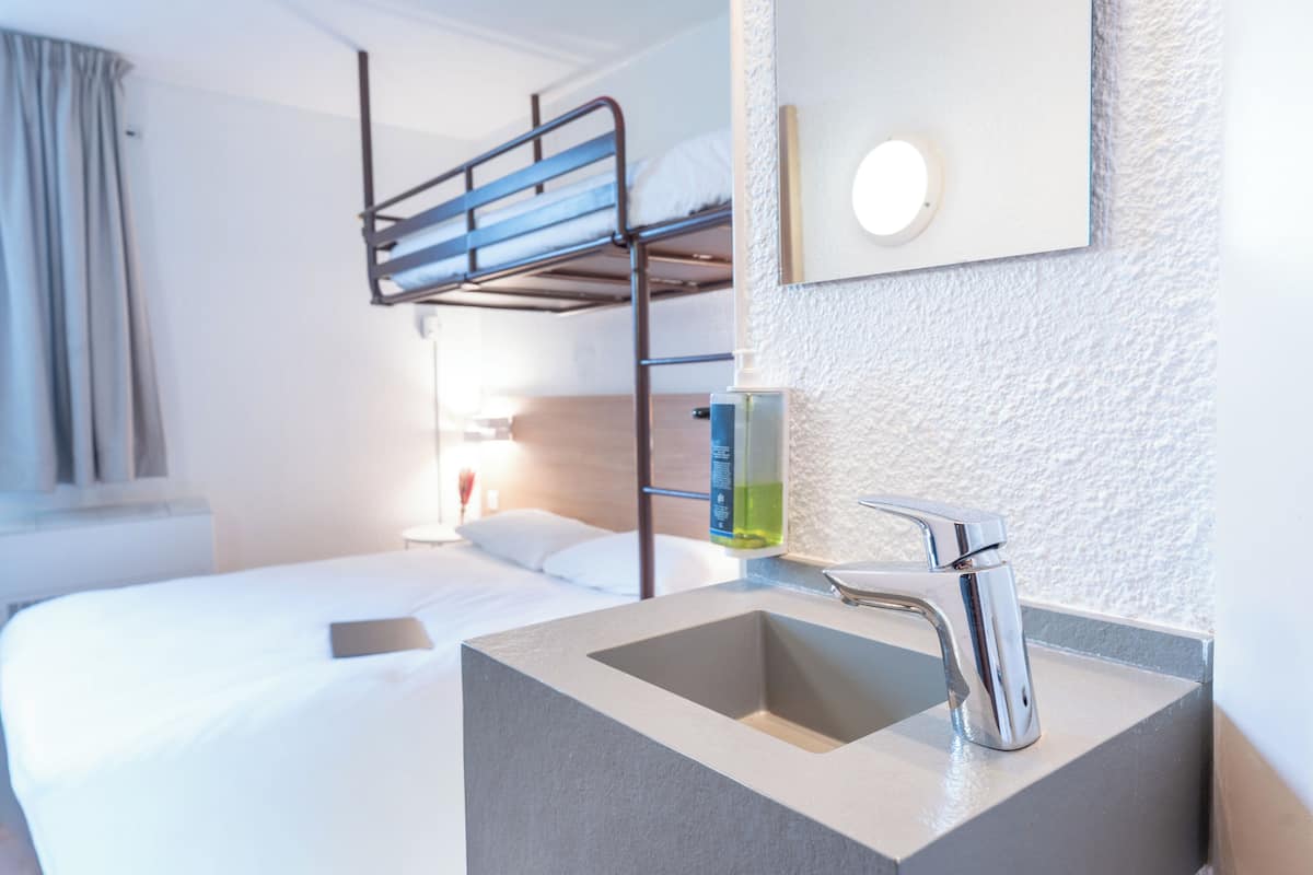A hotel room with a bunk bed, a bedside table with a bottle, a sink, and a mirror.