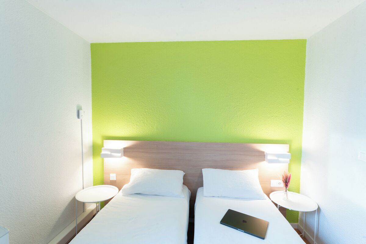 A hotel room with two beds, a green accent wall, bedside lamps, and a laptop on one bed.