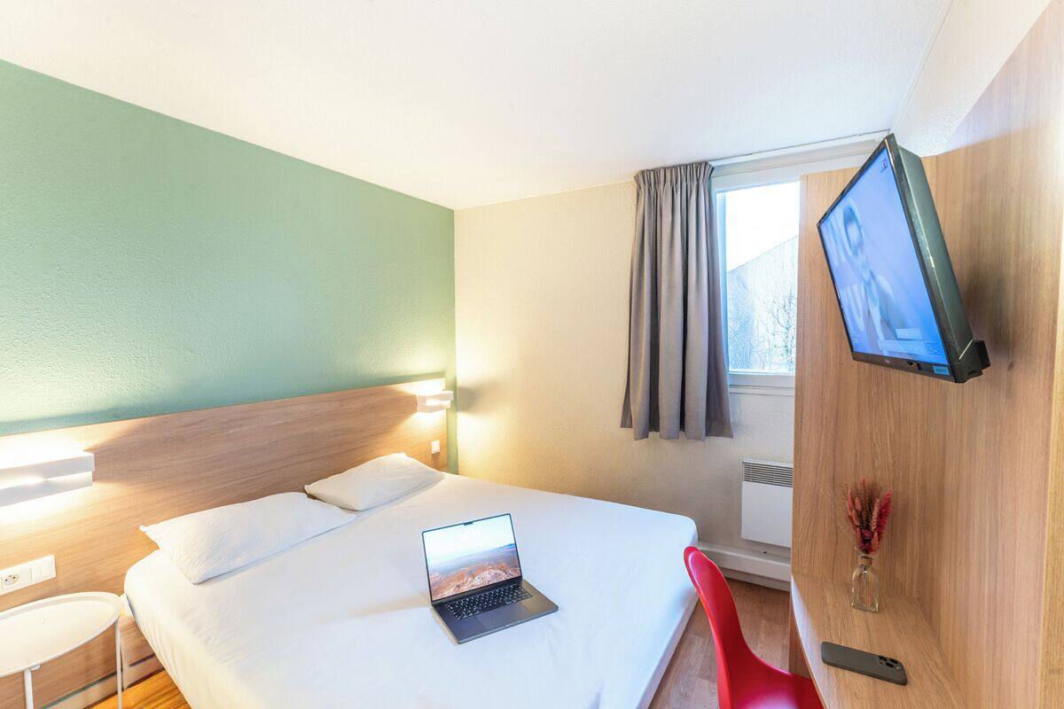 A hotel room with a bed, a laptop, a TV, a red chair, and a vase with flowers.