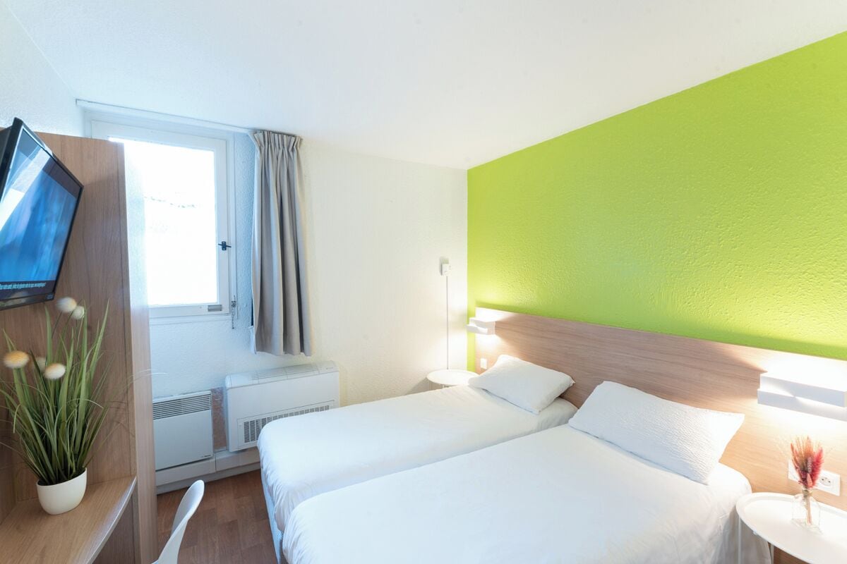 A hotel room with two beds, a TV, a plant, and a window with curtains.