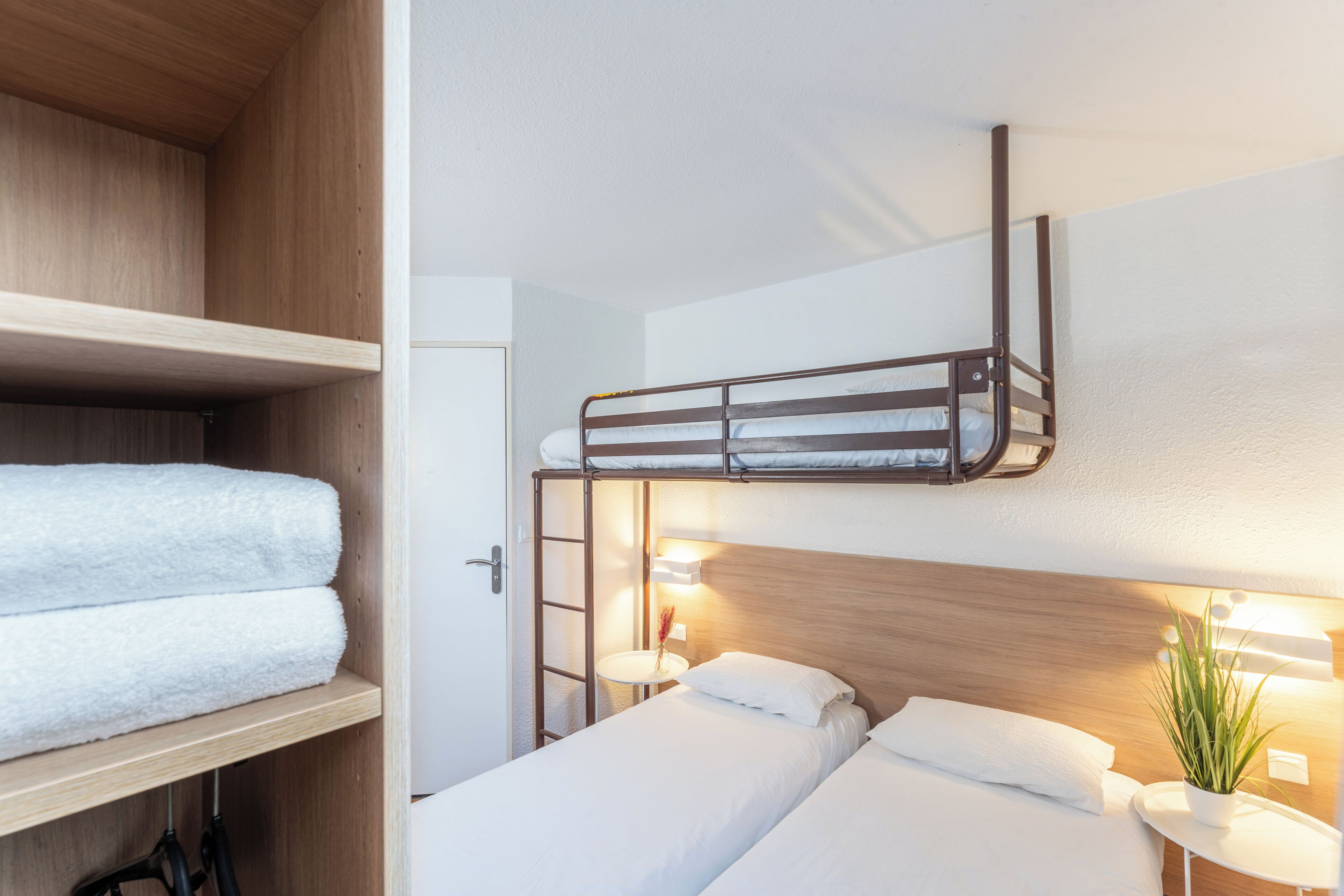 Una habitación con literas, un cabecero de madera, una mesita de noche con una lámpara y una toalla en el estante.