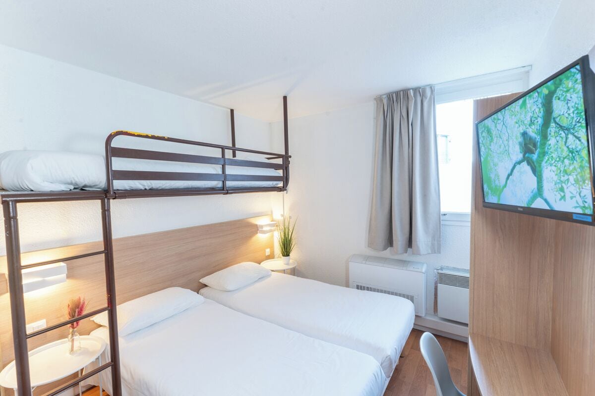 A hotel room with a bunk bed, a flat bed, a TV, a small table with a vase, and a window with curtains.