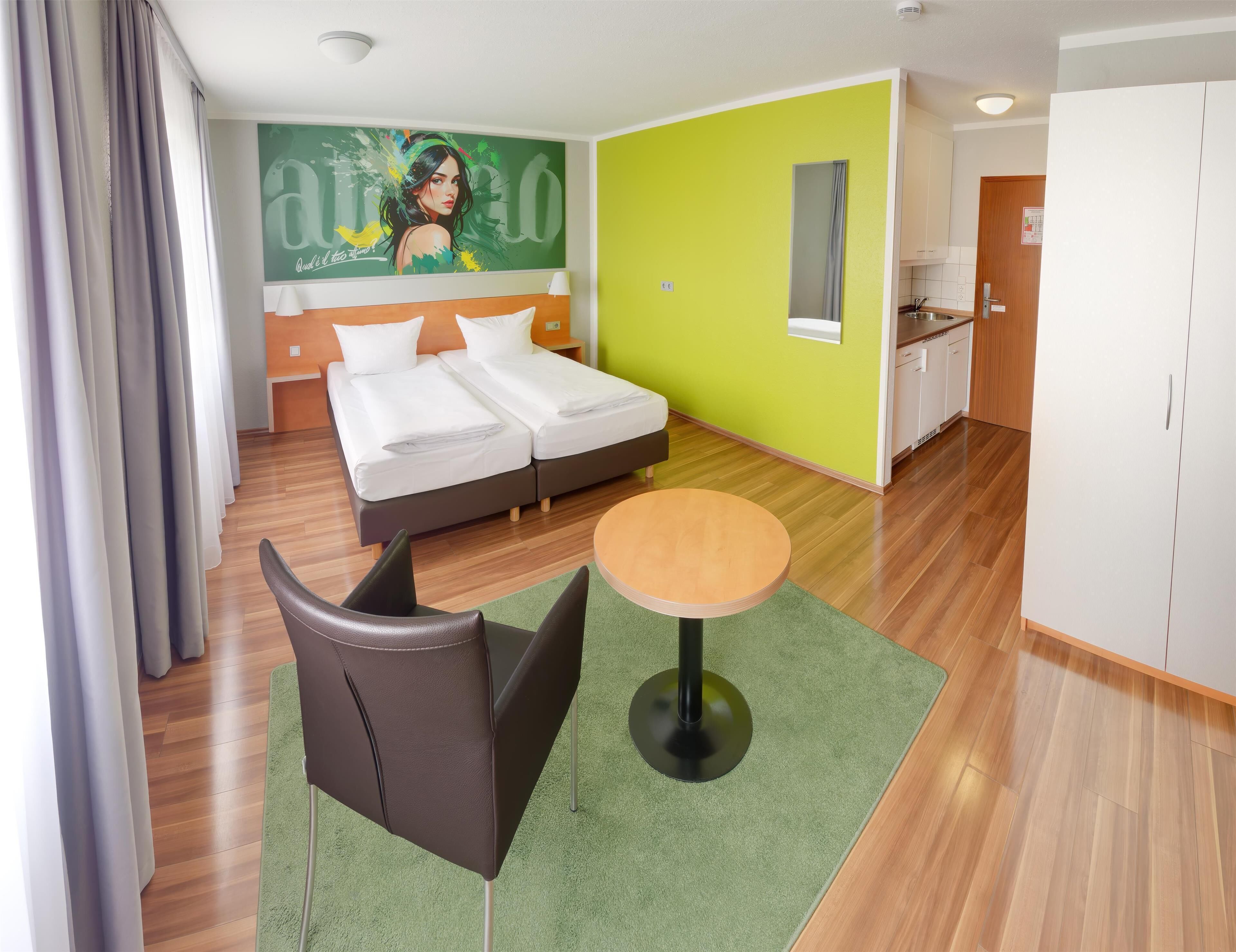A hotel room with a bed, a small table, a chair, a green rug, a painting on the wall, and a kitchen area.