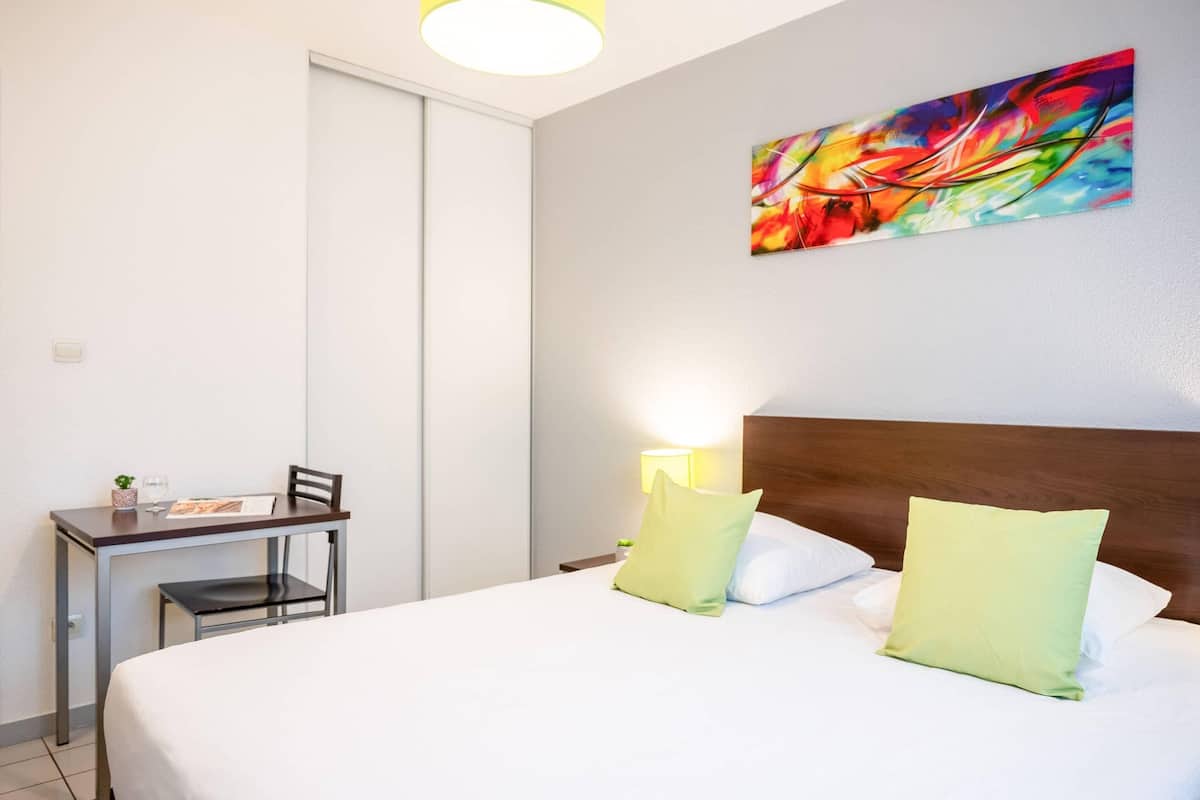 Ein Hotelzimmer mit Bett, Schreibtisch, Stuhl, Lampe und einem farbenfrohen, abstrakten Gemälde an der Wand.