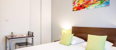Habitación de hotel con cama, escritorio, silla, lámpara y un cuadro abstracto colorido en la pared.