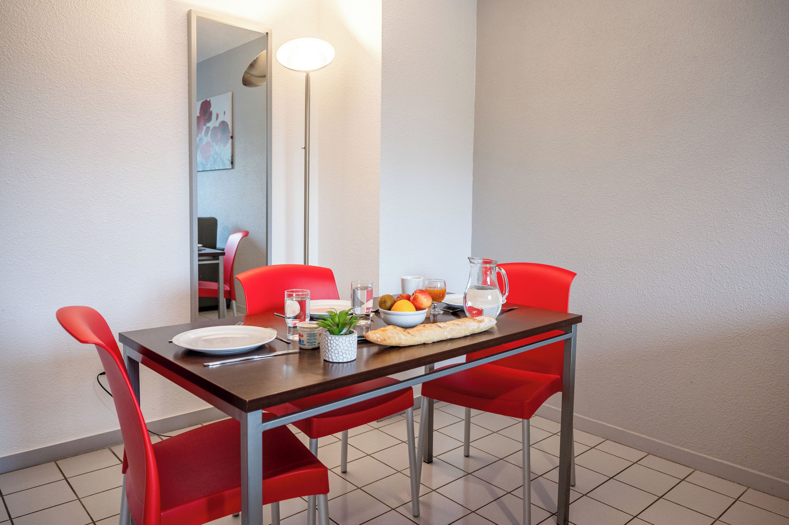 A dining area with a table set for two, red chairs, a floor lamp, and a bowl of fruit.