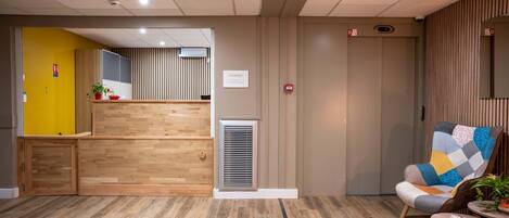 Un área de recepción con mostrador de madera, una planta en maceta y una silla con cojín de parches.