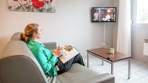 A person sitting on a grey sofa in a room with a television on a stand, a small table with a remote control, and a window with curtains. - Comfort Aparthotel Besancon La City (Besancon)
