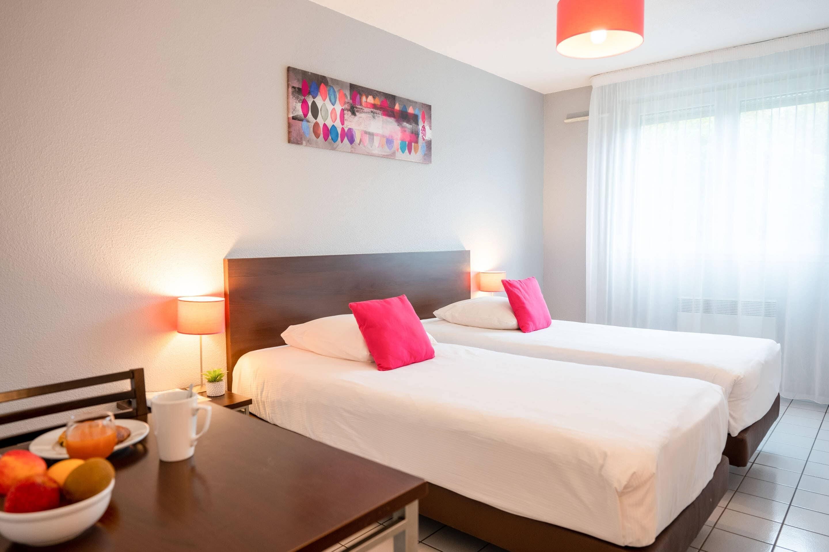 Hotellihuone, jossa on kaksi sänkyä, yöpöytä lampun kanssa, hedelmäkulho ja taulu seinällä.