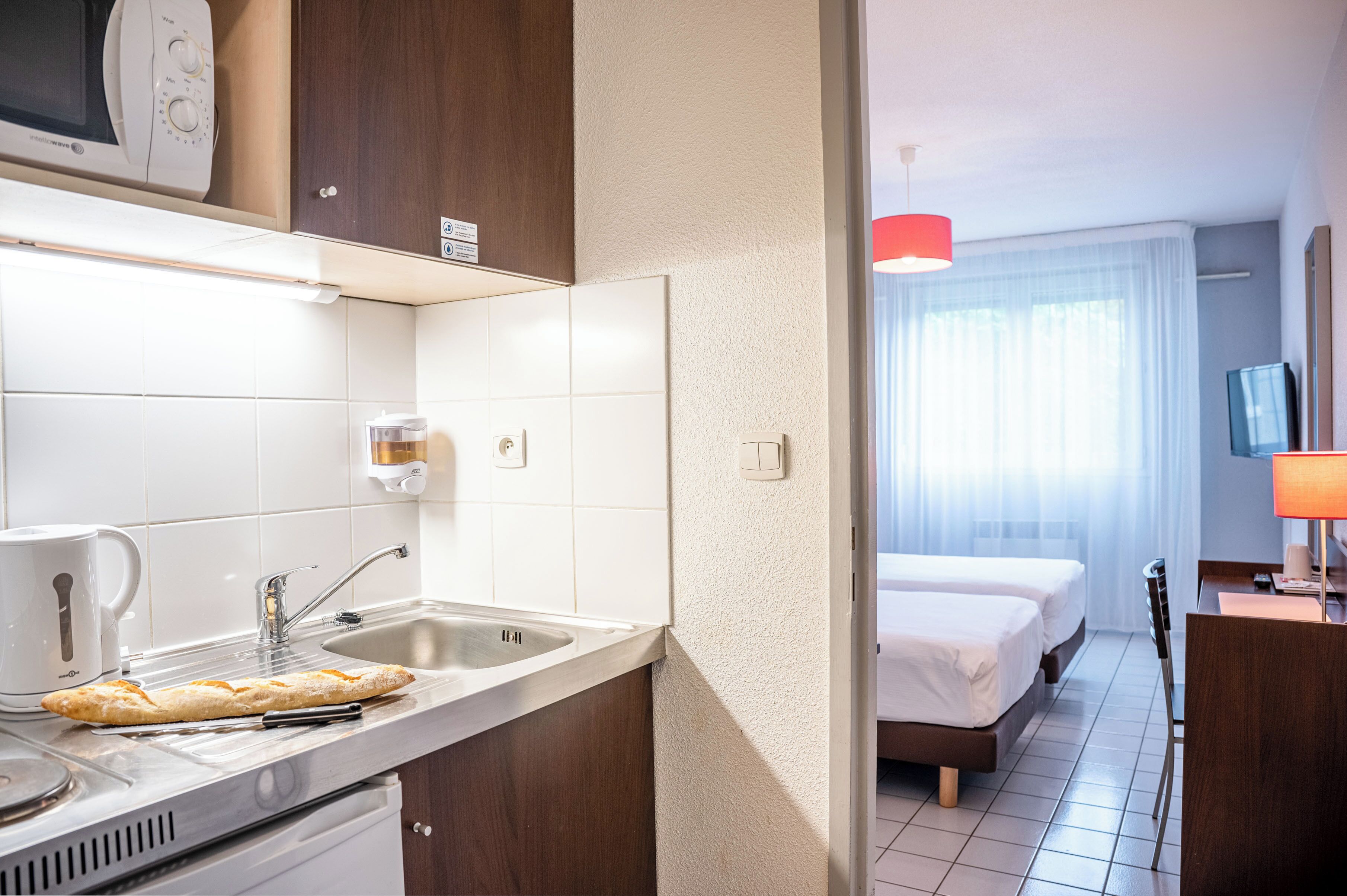 Habitación de hotel moderna con kitchenette, una cama, un televisor y un ventanal con cortinas.