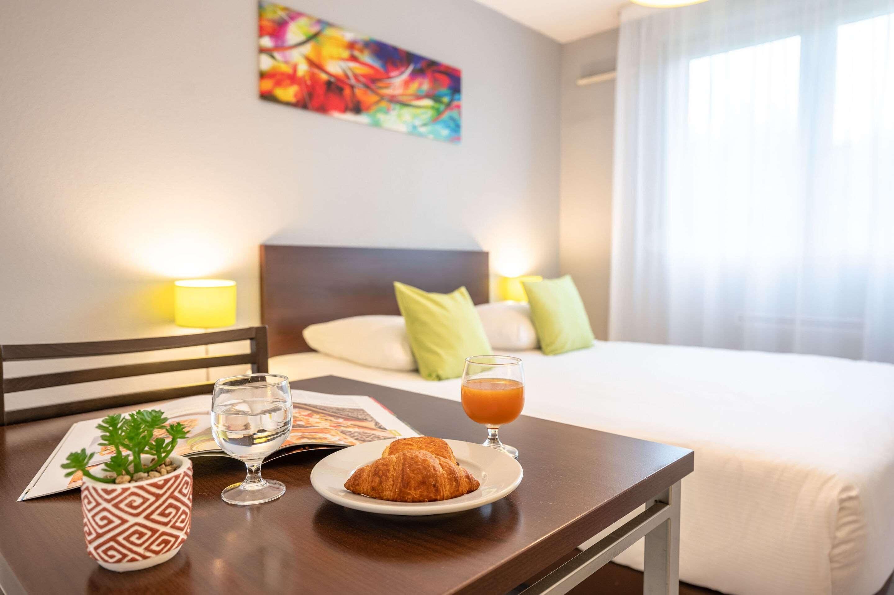 Une chambre d’hôtel avec un lit, une table de chevet sur laquelle se trouve une assiette de croissants, un verre de jus d’orange et une petite plante en pot.