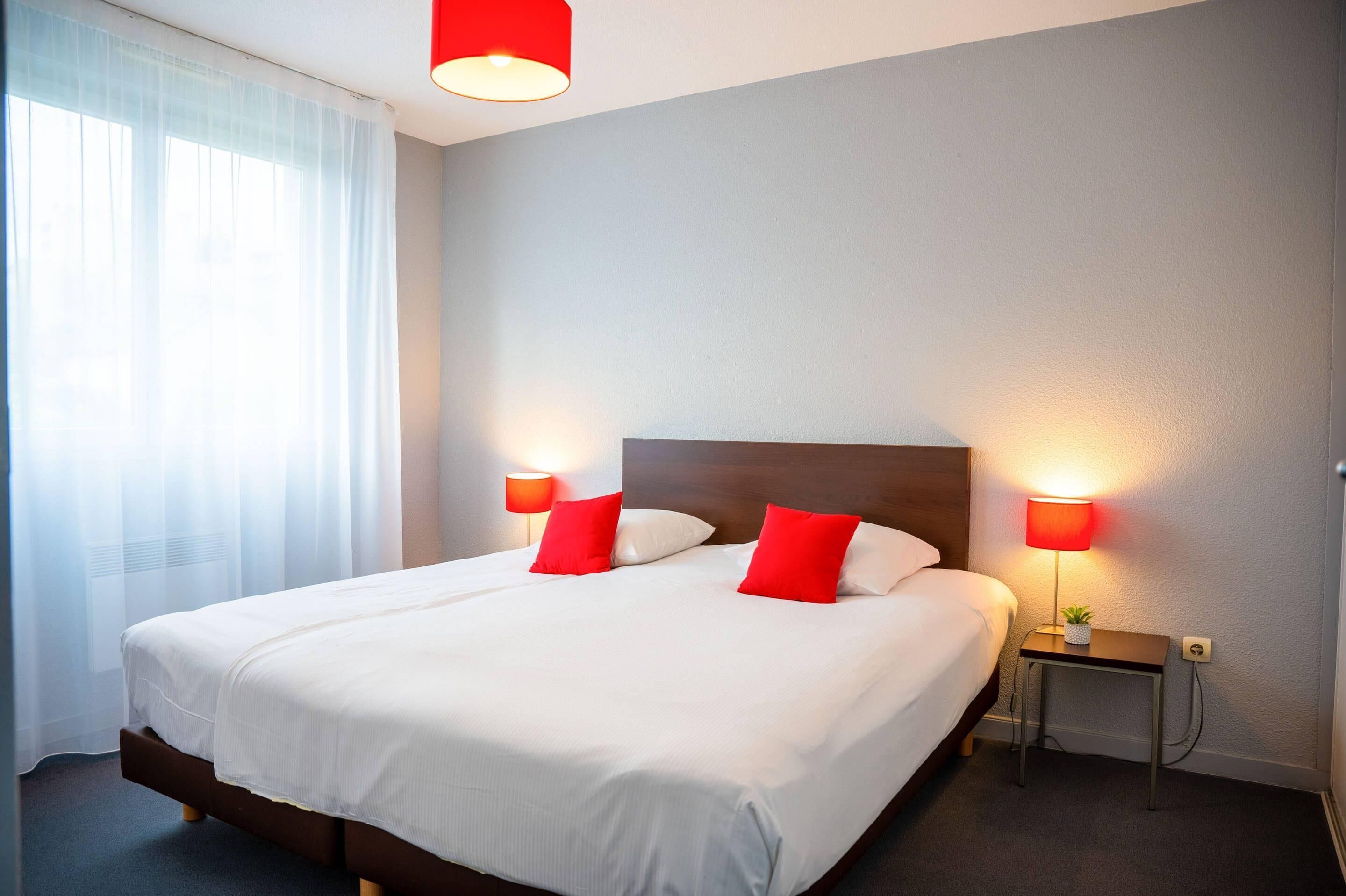 Hotellihuone, jossa on suuri sänky, kaksi yöpöytävalaisinta, pieni pöytä valaisimella ja ikkuna, jossa on ohuet verhot.