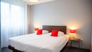 Habitación de hotel con una cama grande, dos lámparas de noche, una mesita con lámpara y una ventana con cortinas translúcidas.
