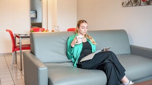 A person sitting on a grey sofa in a room with a red chair, a table with a glass, and a painting on the wall. - Comfort Aparthotel Besancon La City (Besancon)