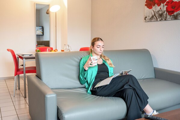 Henkilö istuu harmaalla sohvalla huoneessa, jossa on punainen tuoli, lasilla varustettu pöytä ja seinällä taulu.