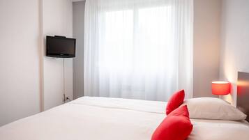Ett hotellrum med en säng, en tv som är monterad på väggen och ett fönster med tunna, genomskinliga gardiner.