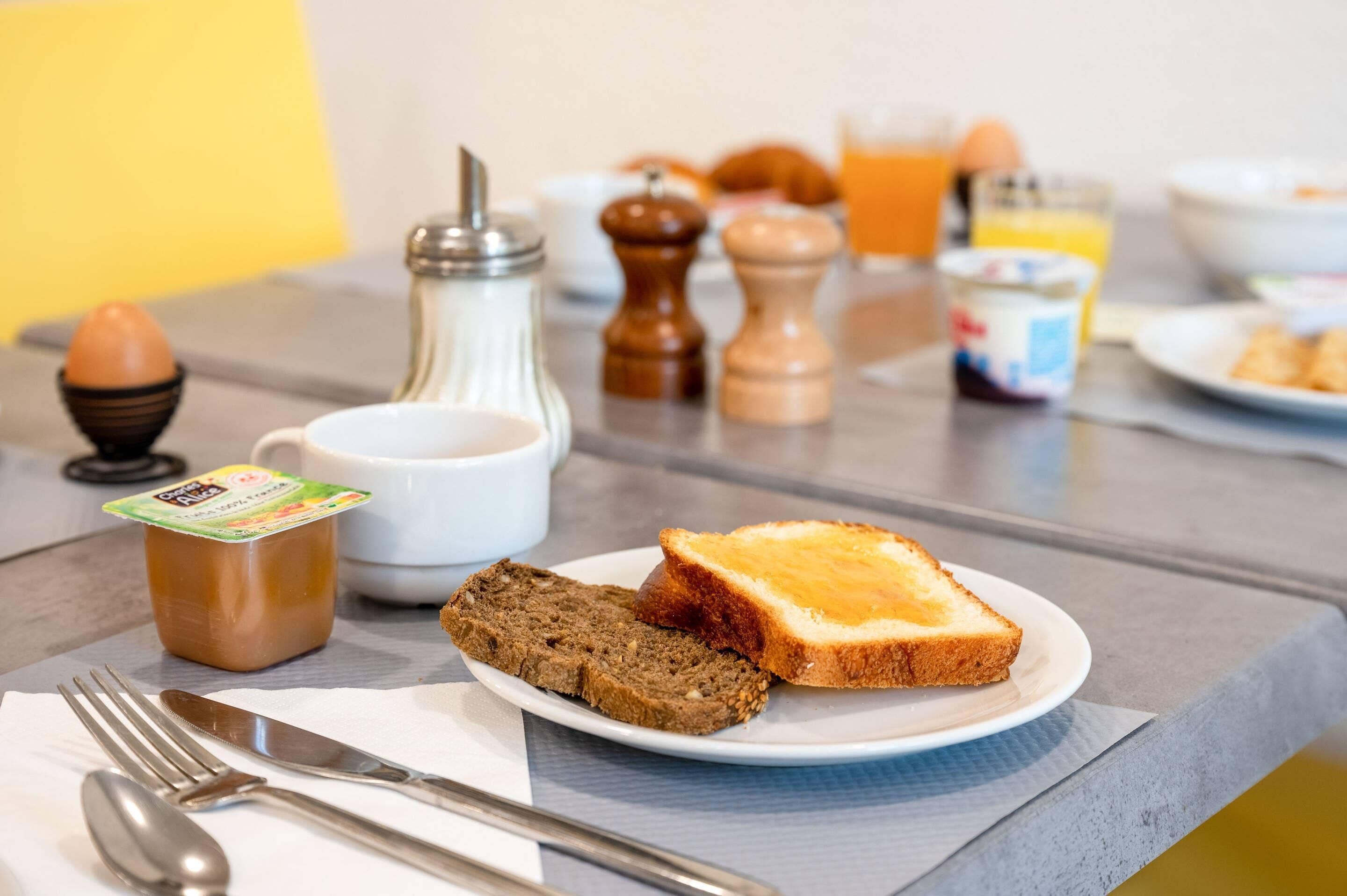 Ein Frühstückstisch mit Toast, Marmelade, einem Salzlöffel, Pfefferstreuern, einer Butterdose, einem Glas Orangensaft und einer Schüssel Müsli.