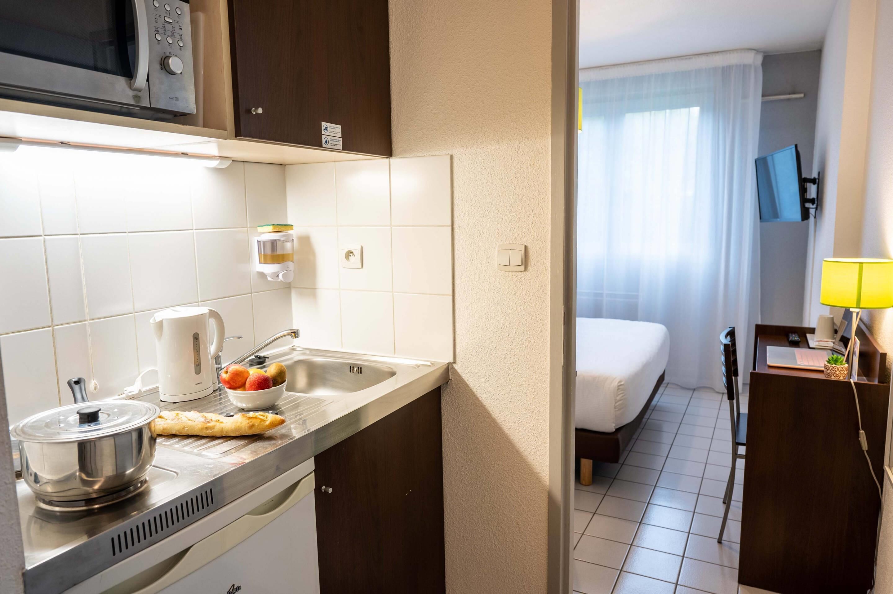 Een kitchenette met een magnetron, waterkoker en spoelbak; een klein zitehoekje met een tafel en stoel; een bed met een televisie; en een bedlampje.