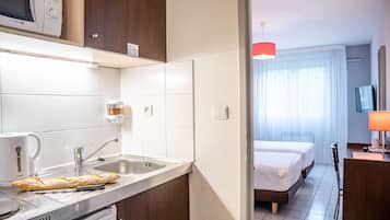 A kitchenette with a coffee maker, a bread loaf, and a toaster. There is a bed with a TV in the adjacent room.