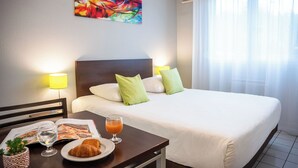 A hotel room with a large bed, two pillows, a bedside table with a magazine, a glass of orange juice, and a croissant. - Comfort Aparthotel Besancon La City (Besancon)