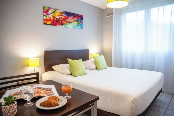 Habitación de hotel con una cama grande, dos almohadas, una mesita de noche con una revista, un vaso de zumo de naranja y un croissant.