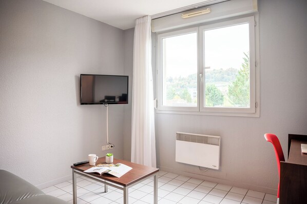 Habitación con un televisor montado en la pared, una mesa con un control remoto, una taza y un libro. Se ve follaje verde por la ventana.