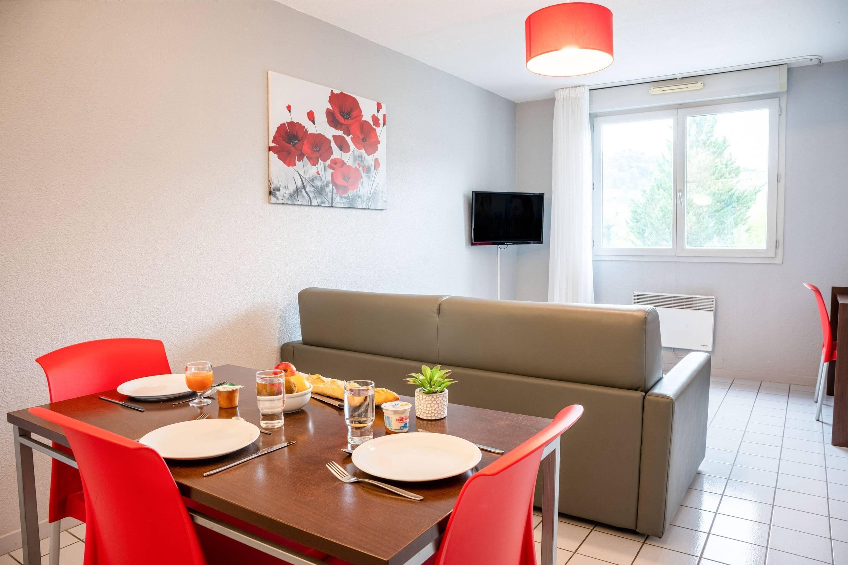 A dining area with a table set for two, a sofa, a TV, and a window with a view of trees.