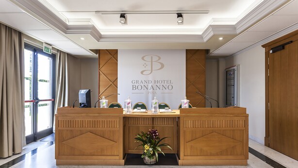 A reception desk at the Grand Hotel Bonanno with a sign above it, a vase with flowers in front, and a view of the outside through the window. - Grand Hotel Bonanno (Pisa)