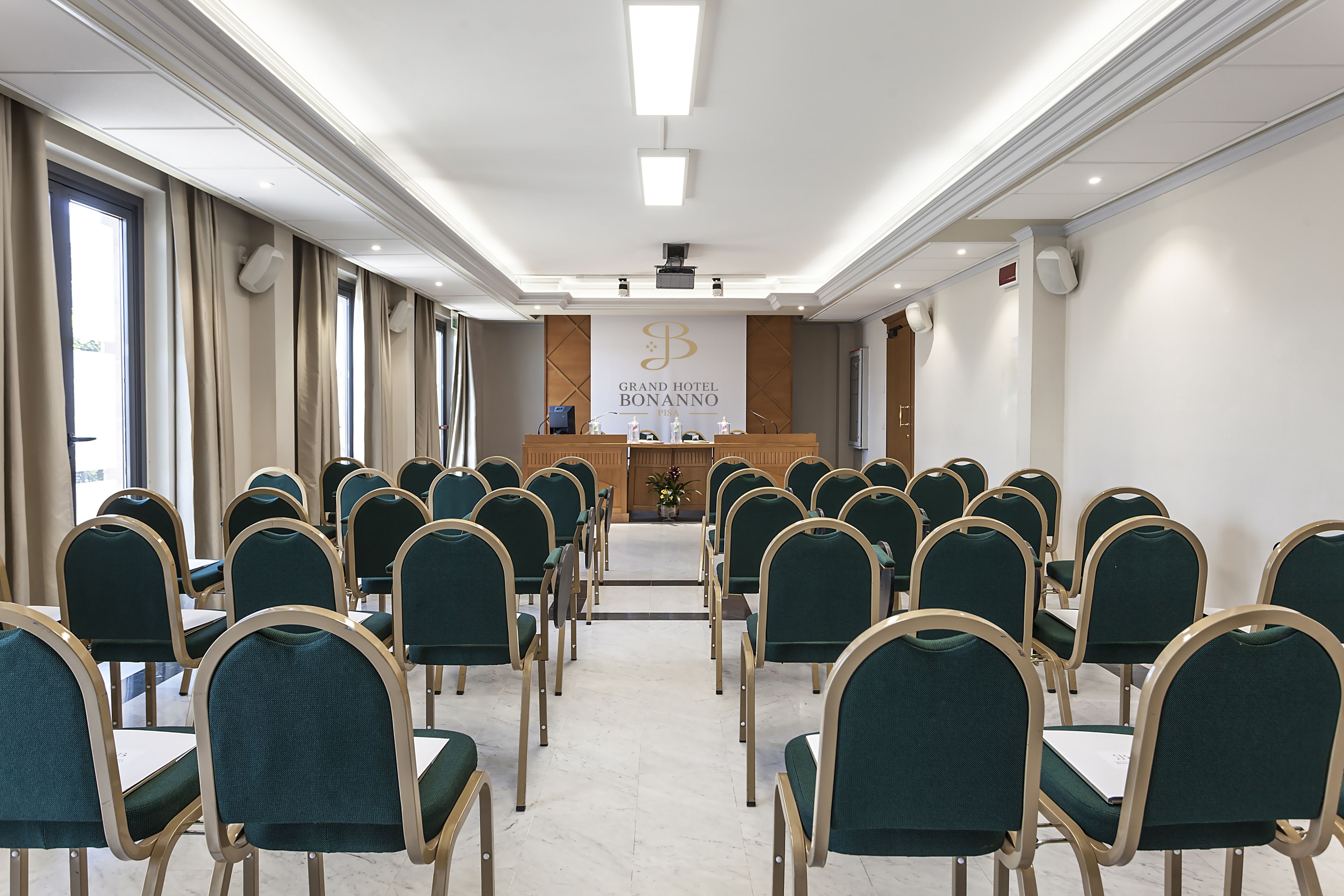 Un salón de conferencias con filas de sillas verdes, un atril y un cartel que dice 'Grand Hotel Bonanno'.