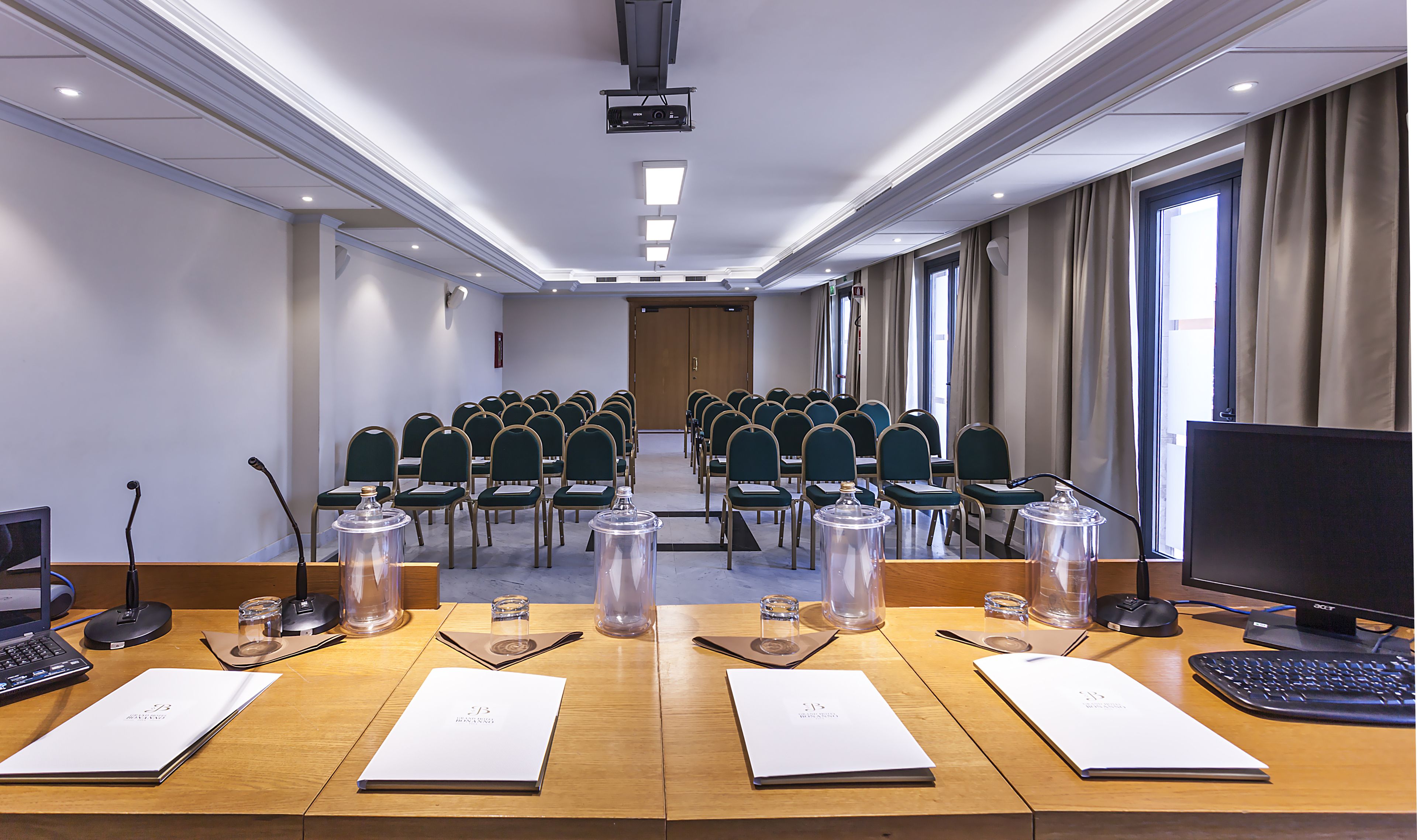 A conference room with rows of chairs, a projector, a large monitor, and a table with documents and a microphone.