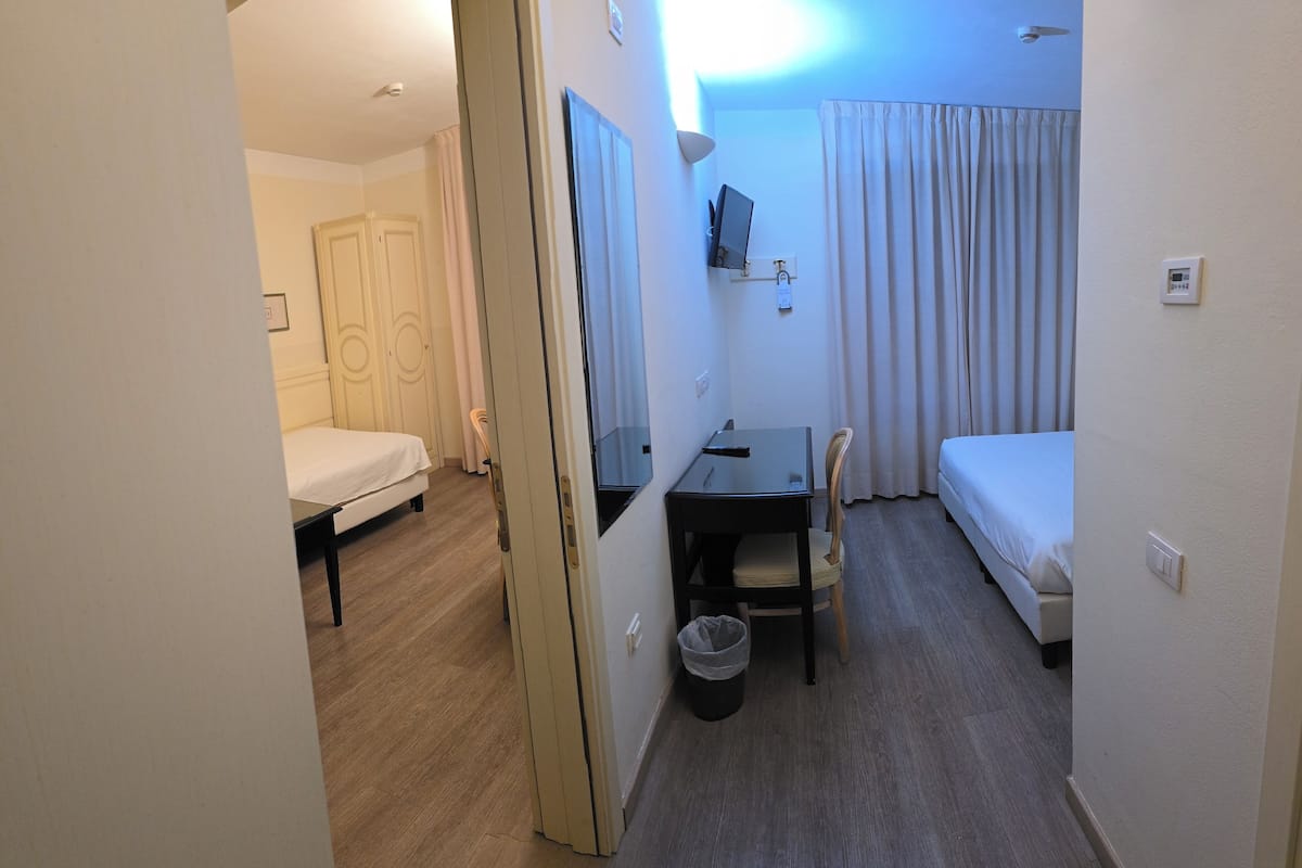 A hotel room with a bed, a desk with a chair, a small table, a TV mounted on the wall, and a trash bin.