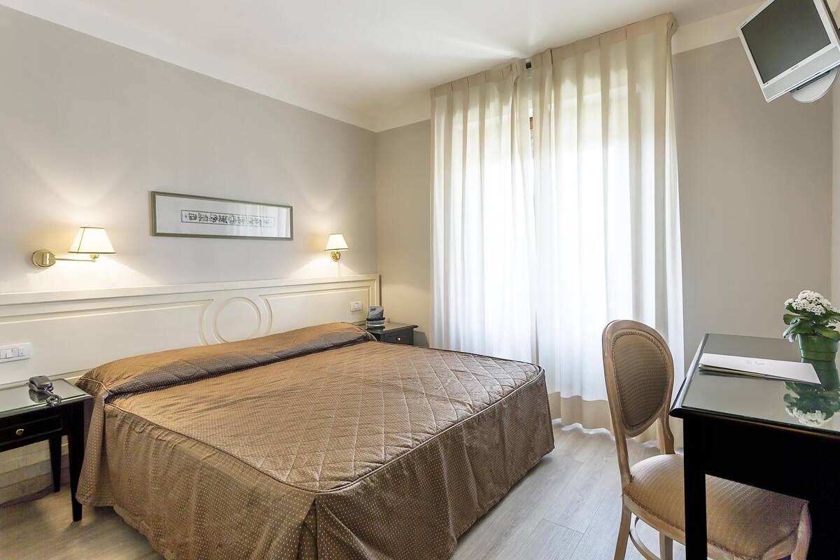 A hotel room with a bed, bedside tables, a desk with a chair, a TV, and curtains.