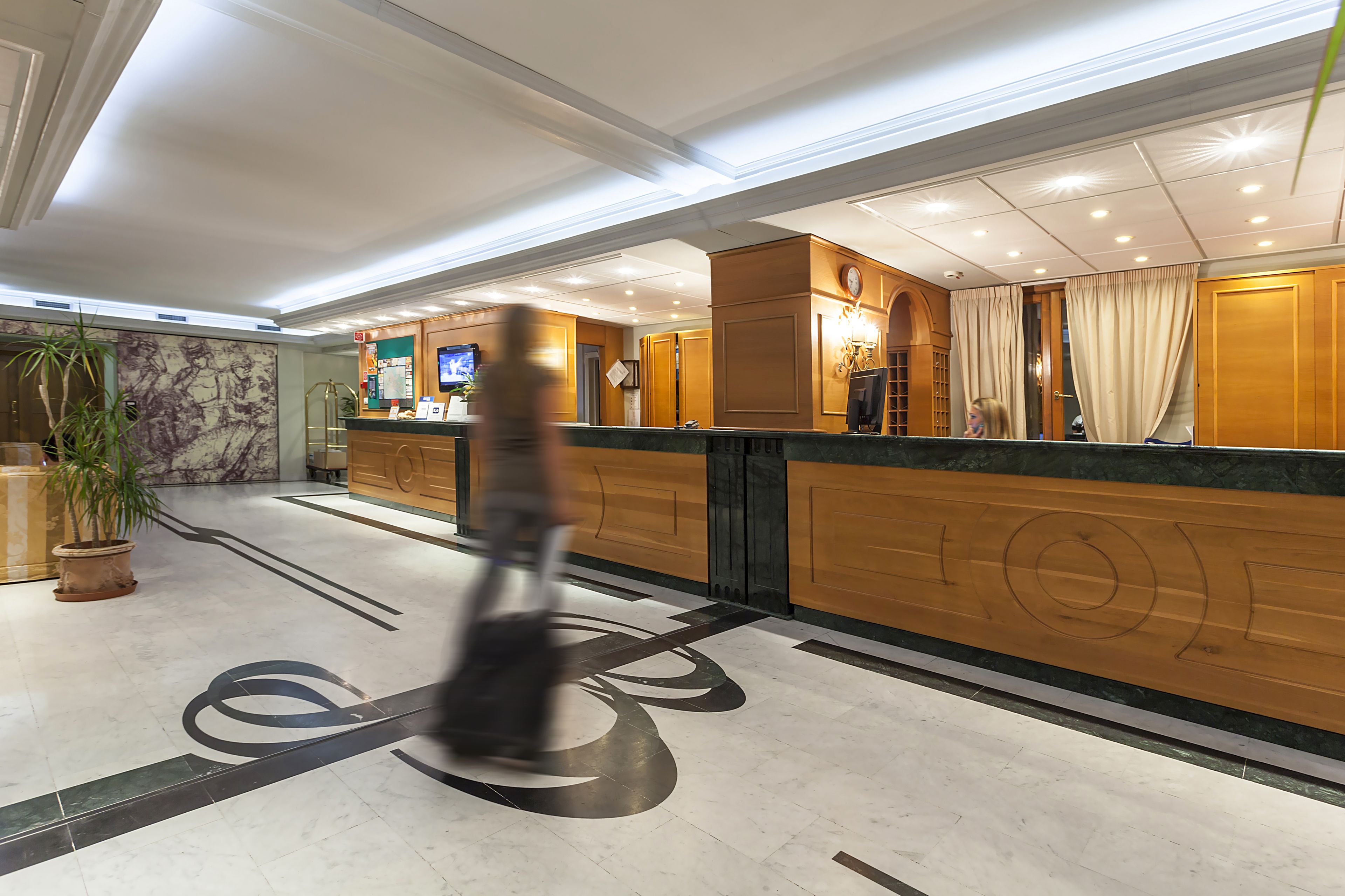 Zona de recepción de un hotel con suelo de mármol, mostrador de madera y una pantalla de televisión.