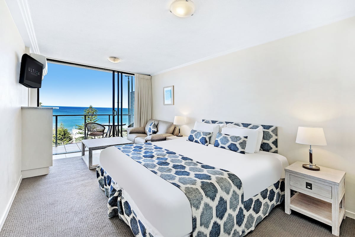 A hotel room with a large bed, a bedside table, a TV, and a balcony with a view of the ocean.