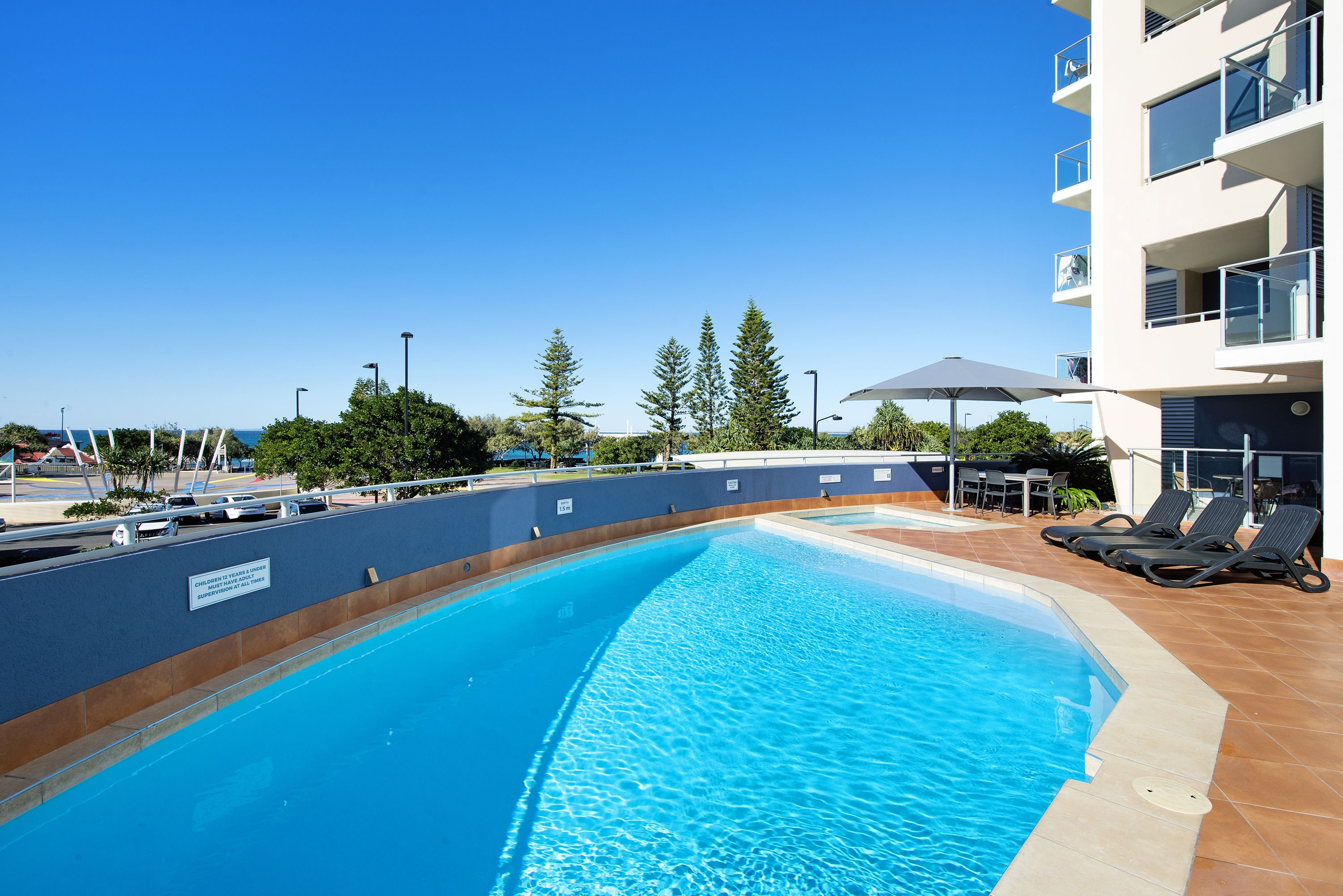 A swimming pool with a clear blue water, surrounded by a tiled deck and a building with balconies.