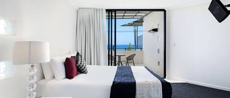 A hotel room with a bed, a bedside table with a lamp, a view of the sea through a glass door, and a small balcony area.
