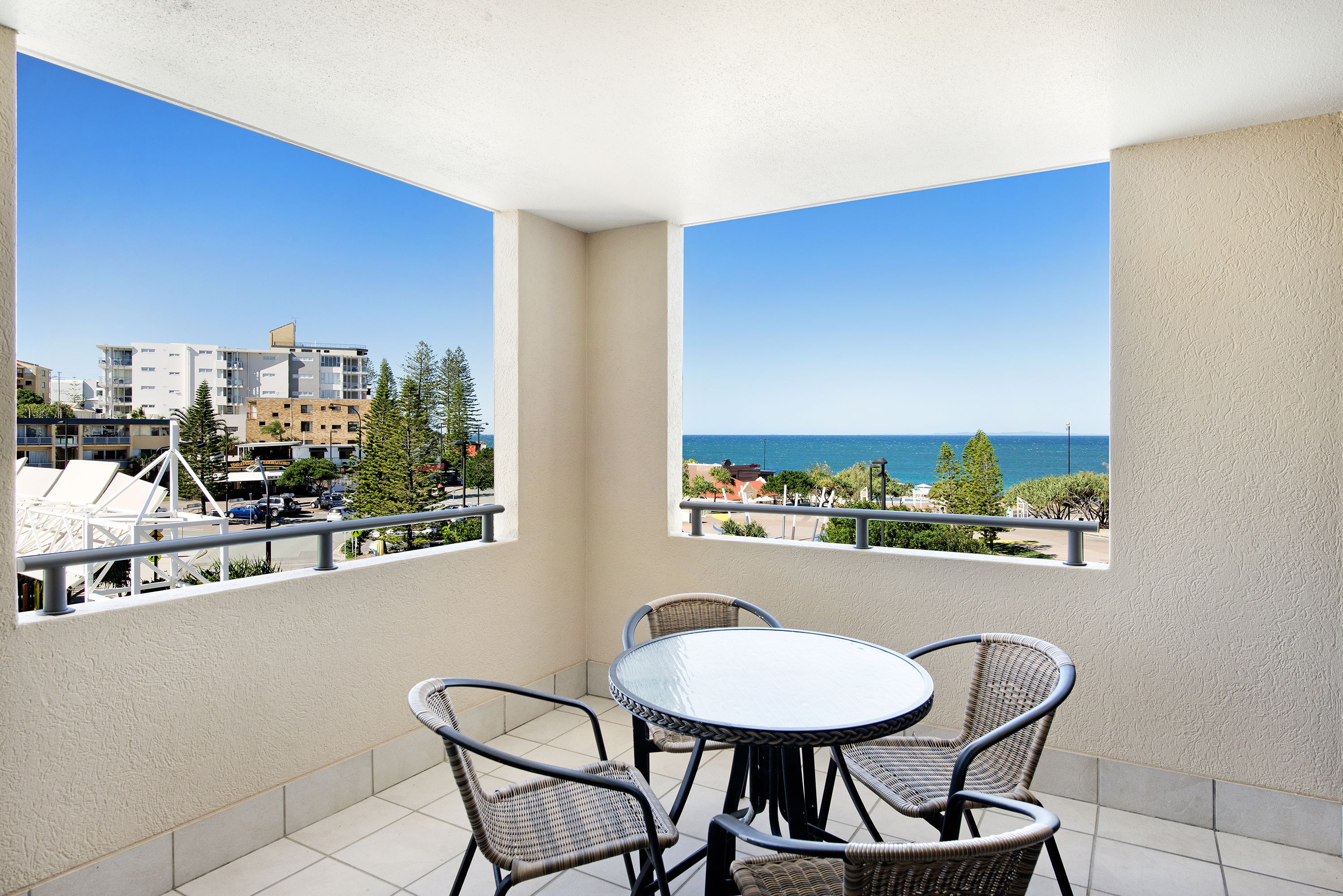 A balcony with a round table and wicker chairs, offering a view of the sea and buildings.