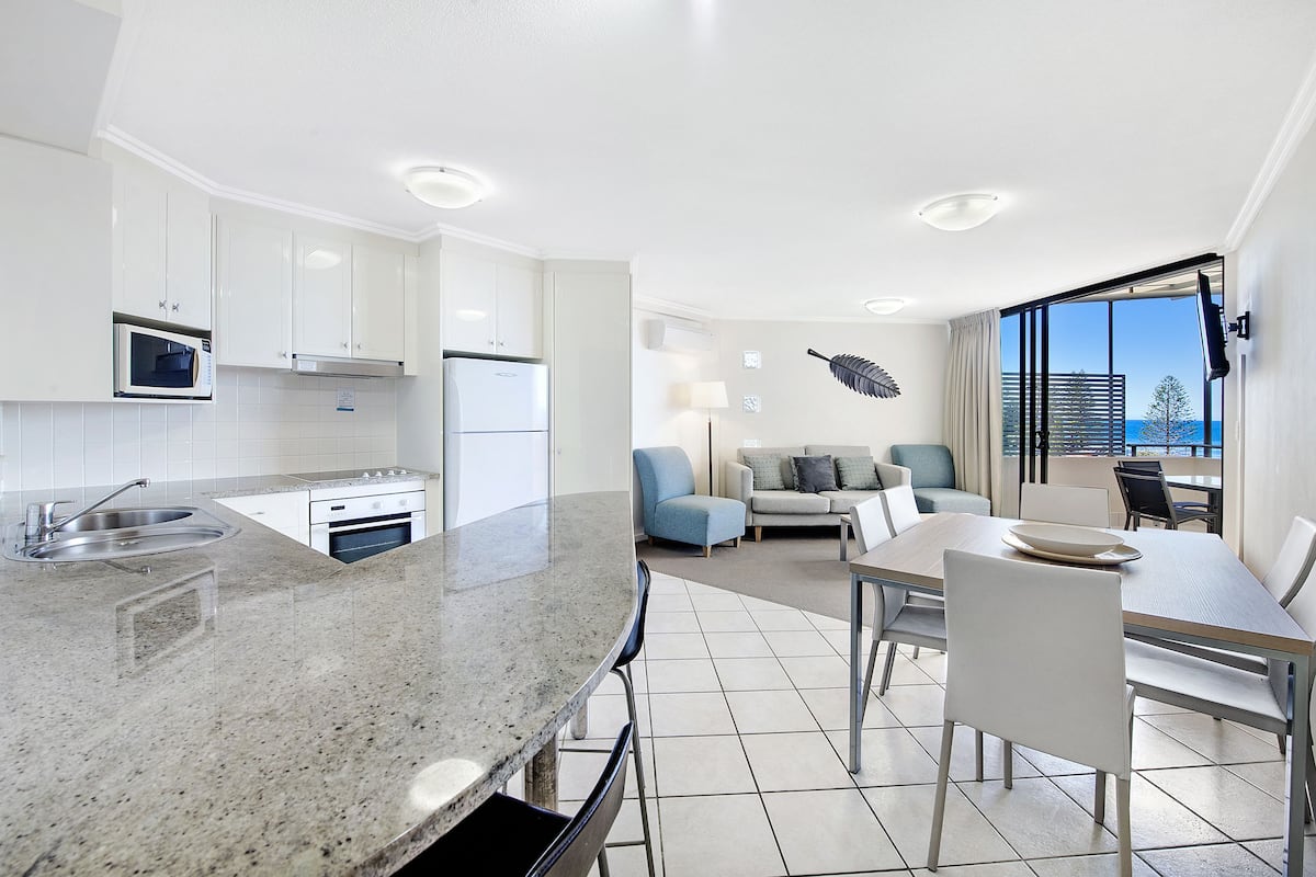 A modern kitchen with white cabinets, a microwave, and a refrigerator. There is a dining area with a table and chairs, and a large window offering a view of the city.