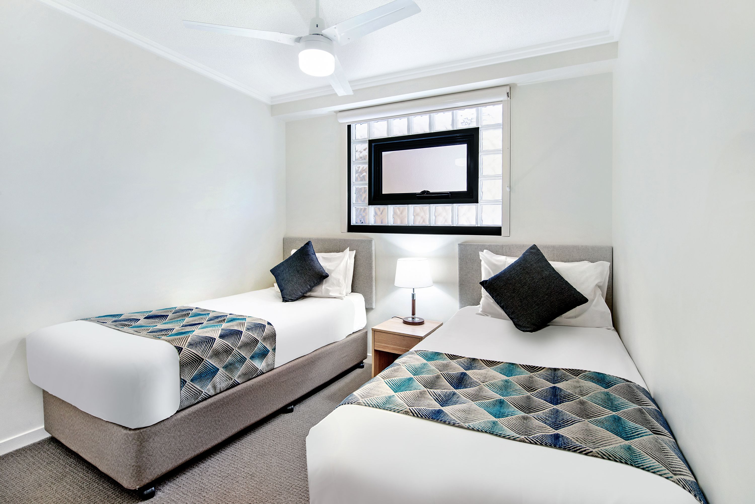 A hotel room with two beds, a TV mounted on the wall, a ceiling fan, and a small bedside table with a lamp.