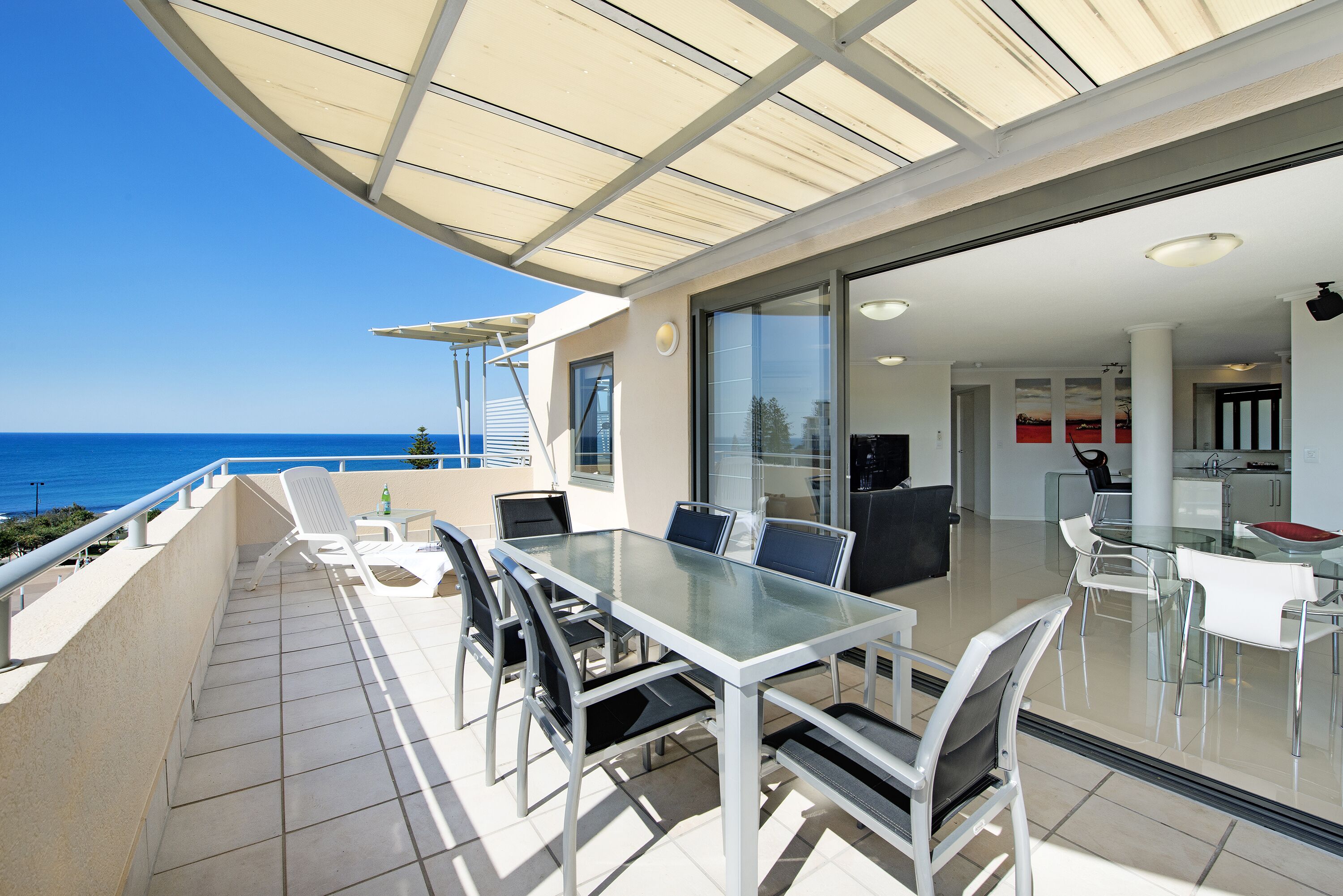 A modern outdoor dining area with a glass table and chairs, a large umbrella, and a view of the sea.