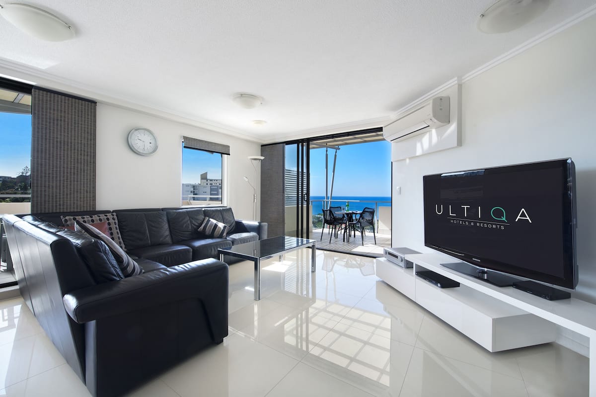 A modern living room with a black leather sofa, a glass coffee table, and a flat-screen TV on a white stand.