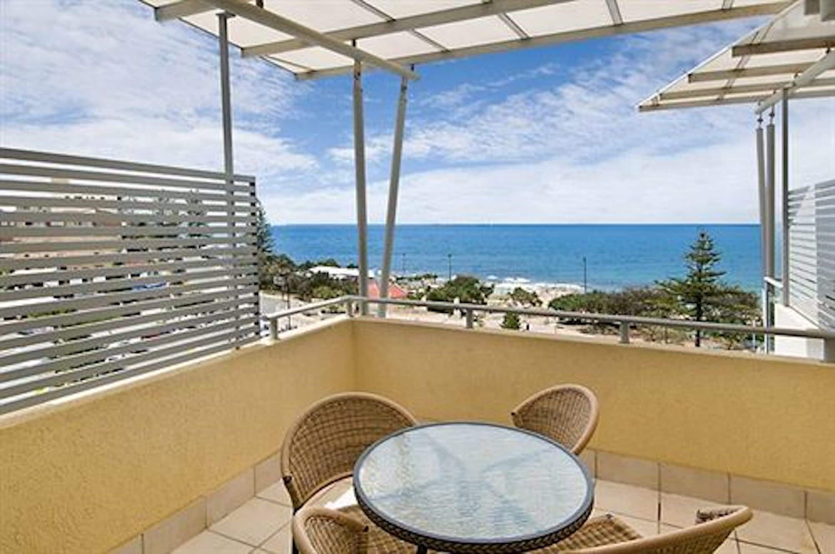 Ein Balkon mit Glastisch und Korbstühlen, der Blick auf einen Strand und den Ozean freigibt.